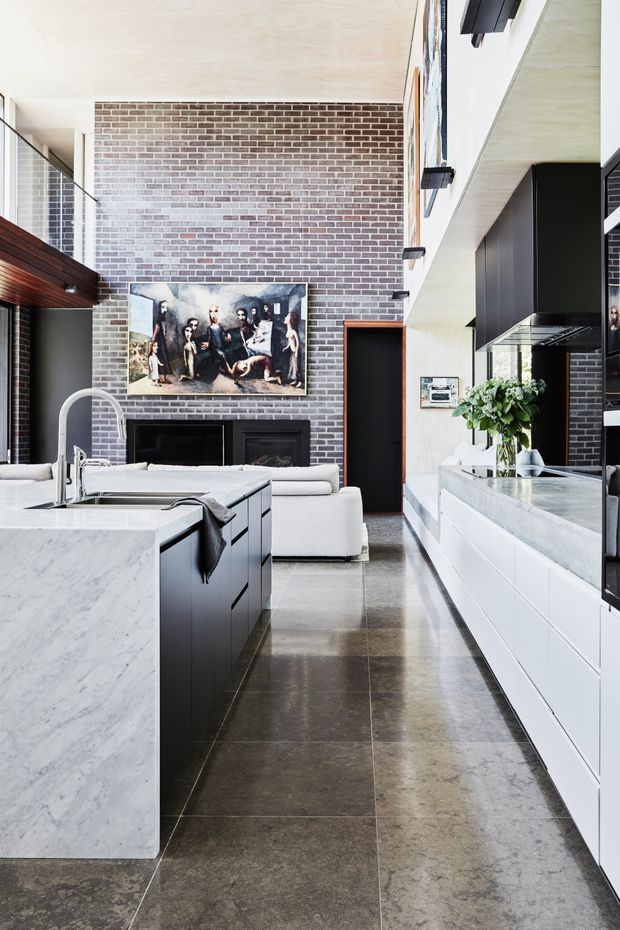 Bright open-plan kitchen and living area with a large marble island, white and dark built-in cabinetry, polished dark stone floor tiles, black rangehood, and a tall exposed brick feature wall. A white sofa and framed artwork sit beyond the kitchen, while timber ceiling detailing and high-level glazing add architectural interest.