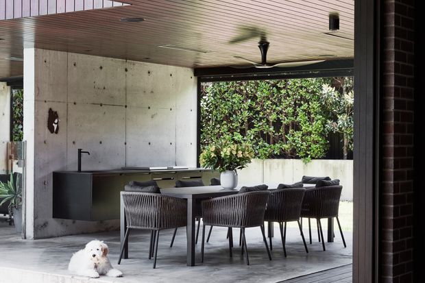 Covered outdoor dining and kitchen area with a long dark dining table, woven dining chairs, and a compact built-in kitchen. The space features exposed concrete walls, a timber-lined ceiling, large open glazing, and a planted courtyard view, creating a refined indoor-outdoor setting.