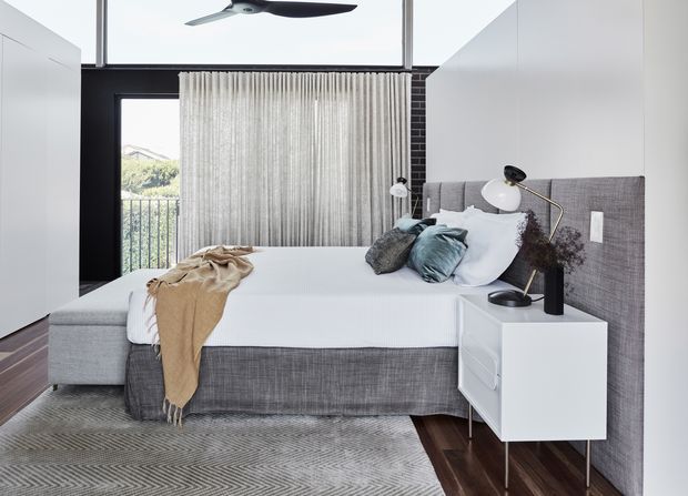 A bright contemporary bedroom with a large upholstered bed, grey textured headboard wall, white floating bedside cabinetry, layered neutral bedding, and a patterned grey rug. Full-height glazing with sheer curtains opens toward a balcony, while a black ceiling fan and dark timber flooring add contrast.