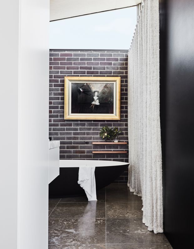 A sophisticated bathroom viewed through a partially open doorway, featuring a freestanding black bathtub, dark brick feature wall, large dark stone-look floor tiles, a sheer textured curtain, and a framed artwork above a narrow plant shelf. The palette is dark, monochromatic, and refined with strong textural contrast.