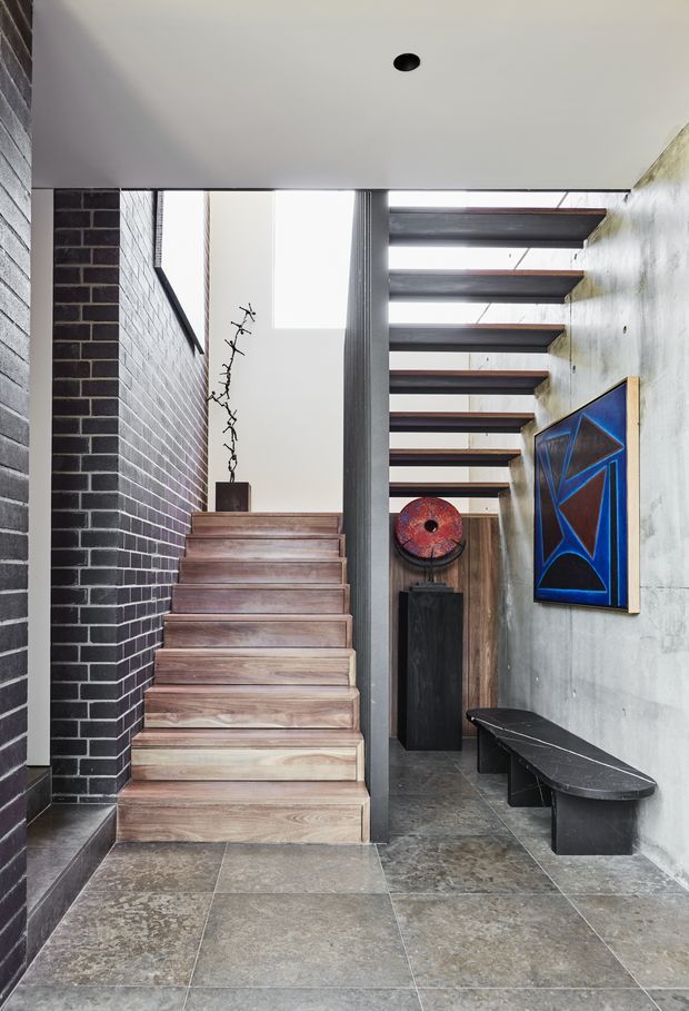 A refined residential entry hall with a central timber staircase, dark brick wall, concrete-look plaster wall, and large stone-look floor tiles. Exposed dark timber ceiling beams span the corridor and a skylit opening brings in natural light. A sculptural bench and abstract artwork create a gallery-like atmosphere.