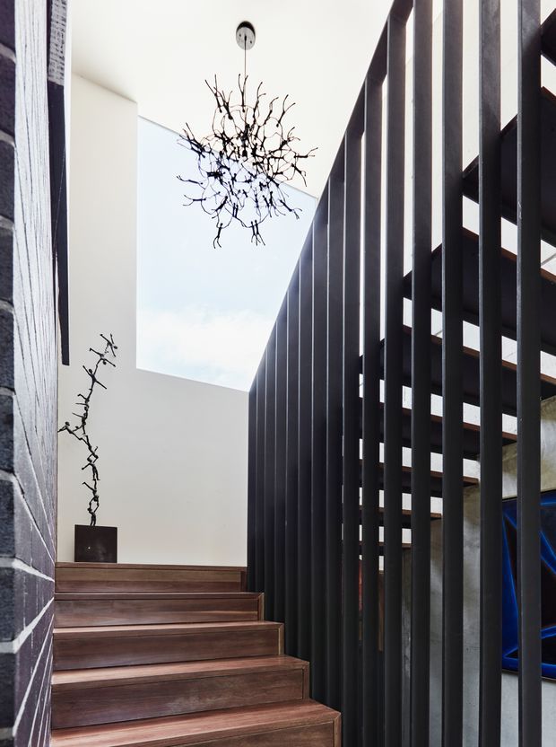 Interior view of a contemporary stairway with broad dark timber treads, a black vertical-bar balustrade, white walls, and a large rectangular skylight or open void above. A sculptural branch-like pendant light hangs over the stair, with a matching branch sculpture on the landing. The space has strong vertical lines and a refined, minimalist architectural character.