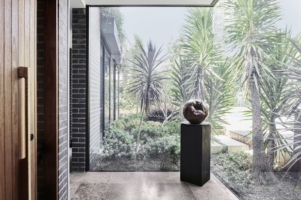 A modern entryway with a tall timber pivot door, dark brick wall, full-height glazing, and a tiled floor. The glazing looks onto a landscaped courtyard featuring sculptural yucca plants and shrubs, while a dark pedestal with a spherical decorative object creates a focal point inside.