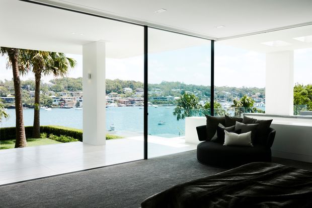 Bright contemporary bedroom overlooking a waterfront through expansive floor-to-ceiling glazing. The room has white walls and large white-format flooring, a dark grey carpet, black lounge chair and bedding, and a minimal built-in window-side ledge. Palm trees, water, and a residential shoreline are visible outside.