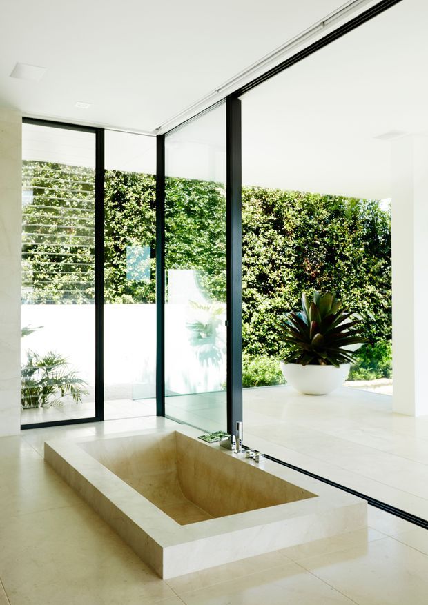 Bright minimalist interior centered on a large square sunken bathtub clad in pale stone, positioned beside full-height black-framed sliding glass doors. The glazing opens toward a private terrace or courtyard with lush hedging and a large potted tropical plant. White walls, pale flooring, and abundant daylight create a clean, open atmosphere.