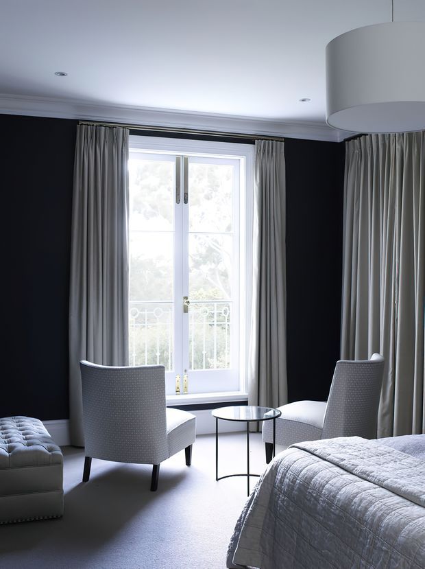 A refined bedroom with charcoal-black walls, a white ceiling with decorative cornice, tall French doors opening to a small balcony, and full-length grey curtains. Two upholstered armchairs face a small round glass-topped table, with a tufted bench and neatly made bed in the foreground. The room has a formal, monochromatic palette and abundant natural light.