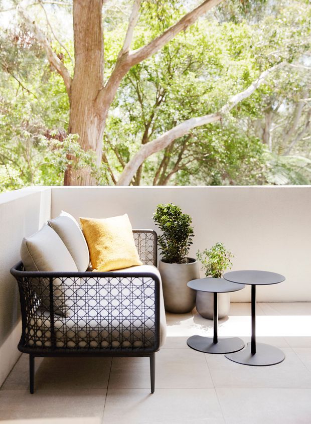 A bright, compact outdoor terrace or balcony furnished with a black woven lounge chair, cream and mustard cushions, two minimalist round black side tables, and potted greenery. The space has pale tiled flooring, smooth light walls, and a low parapet opening onto dense leafy trees, creating a calm, natural setting.