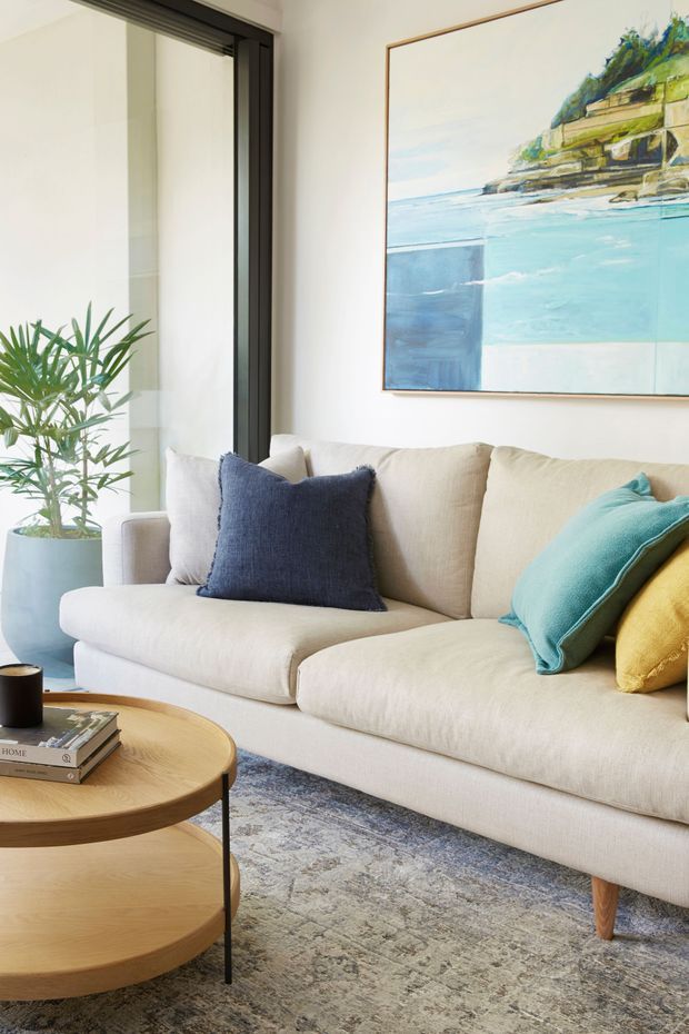 Bright, inviting living room featuring a large cream upholstered sectional sofa with navy, turquoise, and mustard cushions, a round two-tier timber coffee table, a patterned area rug, a potted palm, and a large coastal artwork. Dark-framed glazing or a sliding door adds contrast against the light walls.