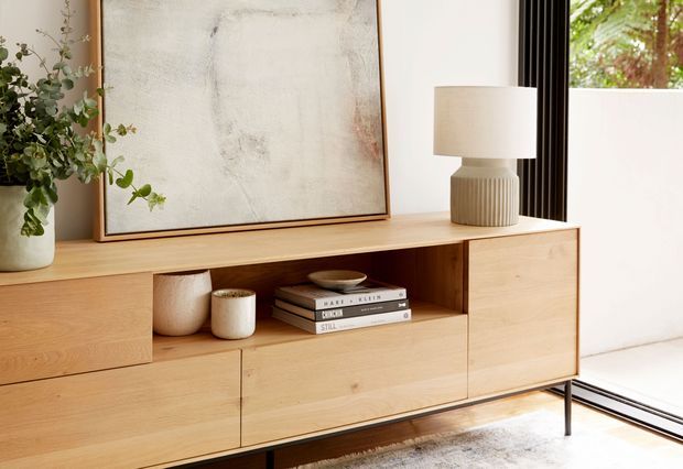 A warm, minimalist living room vignette featuring a low timber sideboard with flat-front cabinetry and an open shelf, styled with books, ceramic vessels, a table lamp, plant, and abstract artwork. Full-height black-framed glazing or a door is visible at right, with a rug beneath the cabinet.