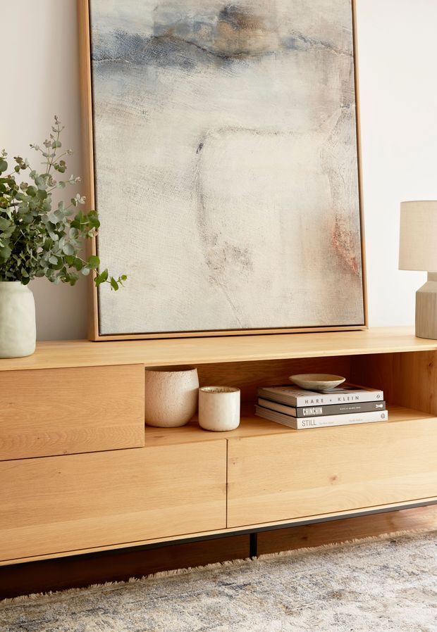 Close-up of a warm, minimalist living-room media console in light timber, with clean-lined drawers and open shelving styled with ceramic vessels and books. A large abstract artwork in a timber frame, leafy plant, and table lamp sit on top, with a neutral textured rug below.