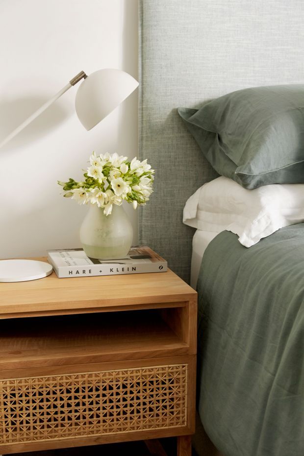 Close-up of a softly styled bedroom corner featuring a timber bedside table with woven rattan-fronted storage, a white wall-mounted reading lamp, a ceramic vase with flowers, and stacked books. The bed has muted sage-green linen bedding with white sheets, positioned against a textured pale green upholstered or fabric wall panel.
