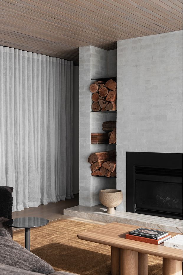 A refined contemporary living room centered on a substantial concrete-block fireplace with open firewood niches and a black inset firebox. Full-height sheer curtains frame the left side, while a timber-lined ceiling, warm timber coffee table, textured neutral rug, and dark upholstered seating create a calm, tactile interior.