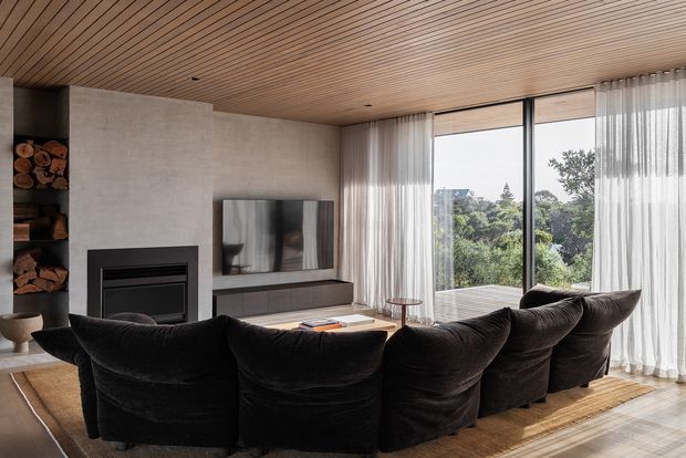 Contemporary living room with a large curved arrangement of dark upholstered lounge chairs facing a wall-mounted television and linear fireplace. The room features a warm slatted timber ceiling, smooth grey concrete or plaster wall surfaces, built-in dark cabinetry, full-height glazing with sheer curtains, and views of greenery outside.