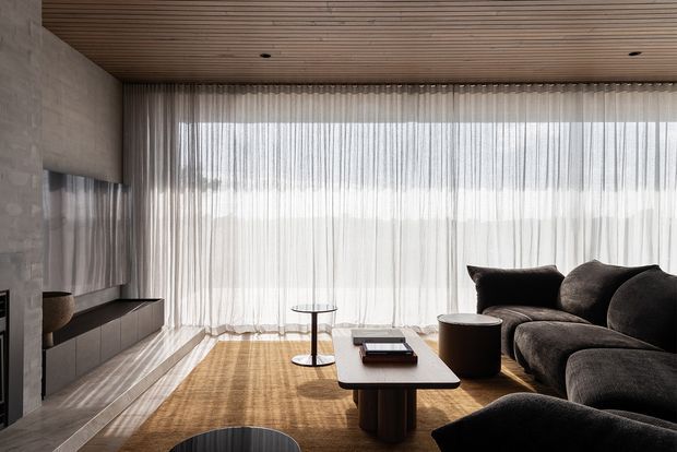 A refined contemporary living room with full-height sheer curtains across expansive glazing, a dark sectional sofa, low rectangular coffee table, slim side tables, and a minimalist built-in media console. The room combines warm timber ceiling slats and flooring with a pale textured feature wall and soft natural light.