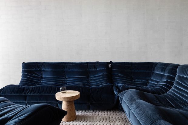 A moody living room with a large deep navy-blue velvet sectional sofa arranged around a small round pedestal side table. The room features a pale textured wall and a light woven rug, creating strong contrast with the dark plush upholstery. The furniture has a sculptural, low-profile form and heavily channelled upholstery.