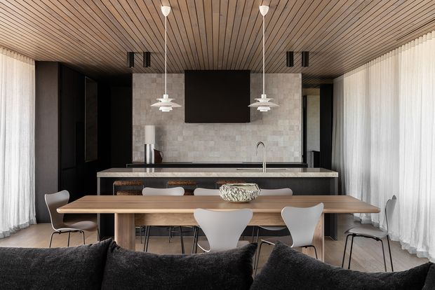 Open-plan contemporary kitchen and dining area viewed from the living space, featuring a long timber dining table, sculptural dining chairs, a dark kitchen with island seating, a pale stone backsplash and benchtop, pendant lights, full-height sheer curtains, and a slatted timber ceiling.