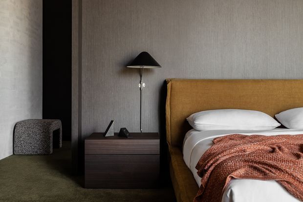 A moody contemporary bedroom featuring a large upholstered mustard-brown headboard, white bedding, and a rust-toned textured throw. A dark wood bedside cabinet with a slim black floor lamp sits against vertically textured dark grey wall panelling, while a muted green carpet and a sculptural upholstered stool add softness.