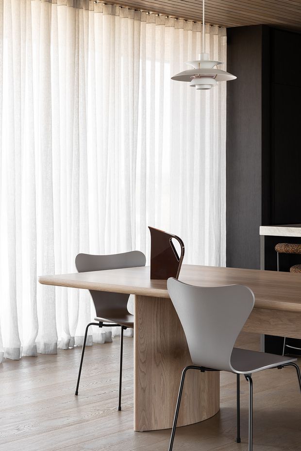 A refined dining area with a light timber dining table, sculptural grey chairs, sheer full-height curtains, and a distinctive suspended pendant light. The room features pale timber flooring and a dark built-in wall or cabinetry contrasting with the bright, softly lit dining space.
