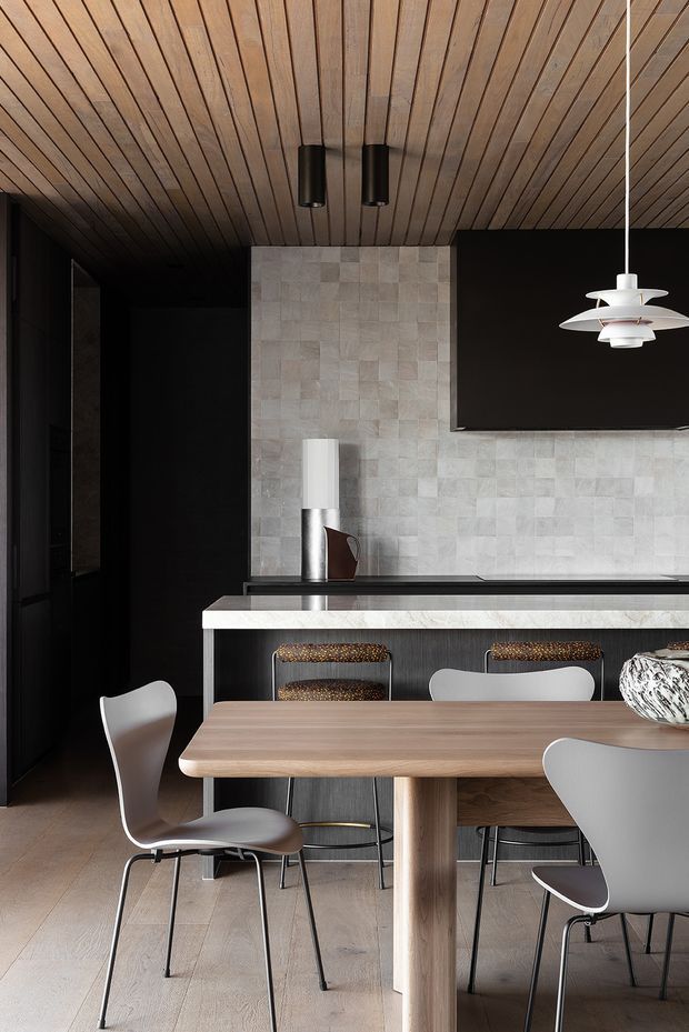 A refined open-plan kitchen and dining area with a warm timber slatted ceiling, pale timber flooring, a large timber dining table, sculptural modern dining chairs, dark cabinetry, a pale textured stone-tile backsplash, and a black kitchen island. Layered pendant and ceiling lighting add a contemporary, high-contrast feel.