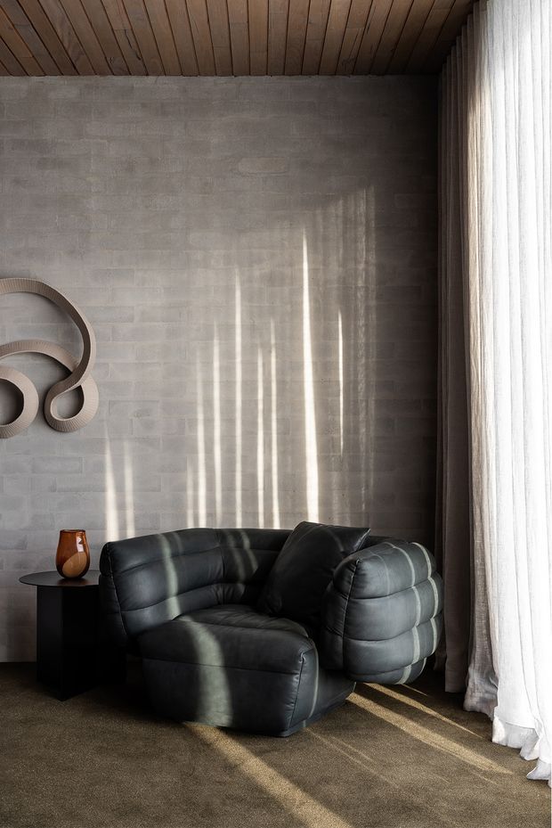 A moody, intimate living room corner featuring a sculptural black leather lounge chair, a small black side table with a ceramic vase, and abstract wall art. The room has a grey concrete block wall, warm timber-lined ceiling, full-height sheer curtains, and soft sunlight casting vertical shadows across the wall, chair, and carpet.