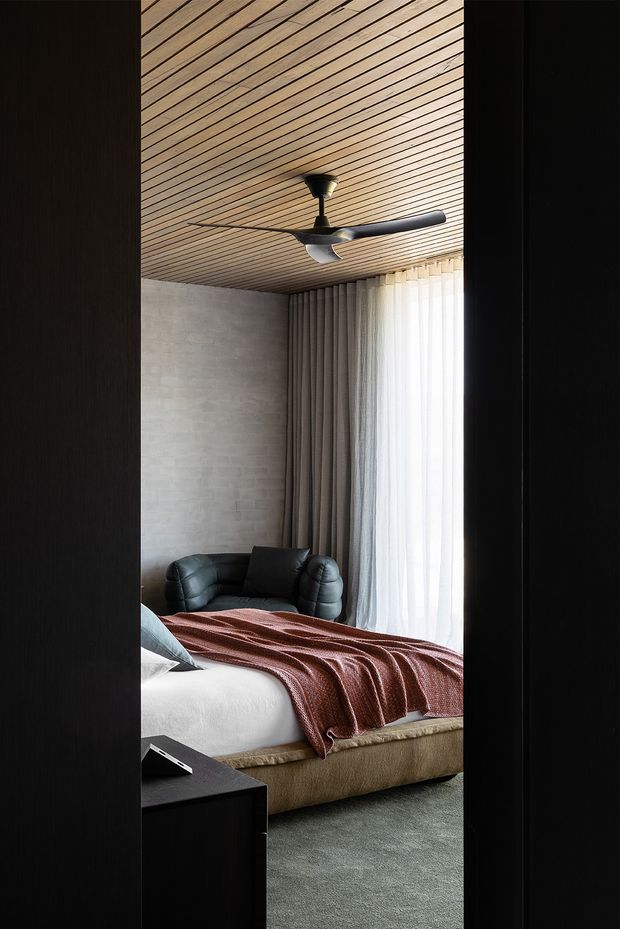 A refined modern bedroom viewed through a dark doorway, featuring a low upholstered bed with a textured rust-red throw, black leather lounge chair, full-height sheer curtains, a pale concrete or masonry feature wall, and a warm timber slatted ceiling with a contemporary ceiling fan. The room has a restrained, tactile palette and strong vertical daylight.