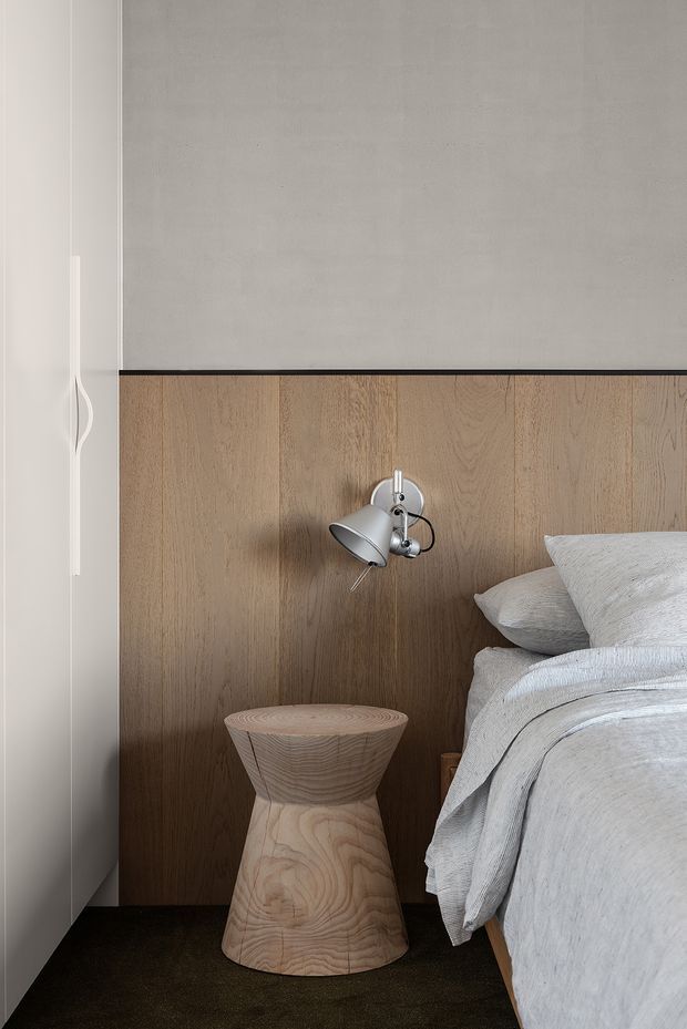Close-up of a calm, minimalist bedroom corner featuring a bed with light grey textured bedding, a sculptural light timber bedside stool, wall-mounted chrome reading light, dark carpet, and a full-height timber wall panel behind the bed. White built-in cabinetry is visible at the left.