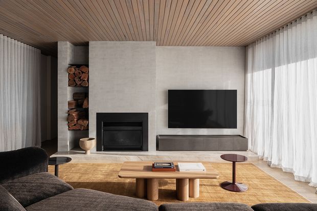 Contemporary living room with a broad concrete fireplace wall, integrated log storage, black fireplace insert and wall-mounted television. Full-height sheer curtains frame large glazing, while a slatted timber ceiling, timber flooring and a low sculptural timber coffee table add warmth. A dark curved sectional sofa and restrained furnishings create a refined, calm interior.