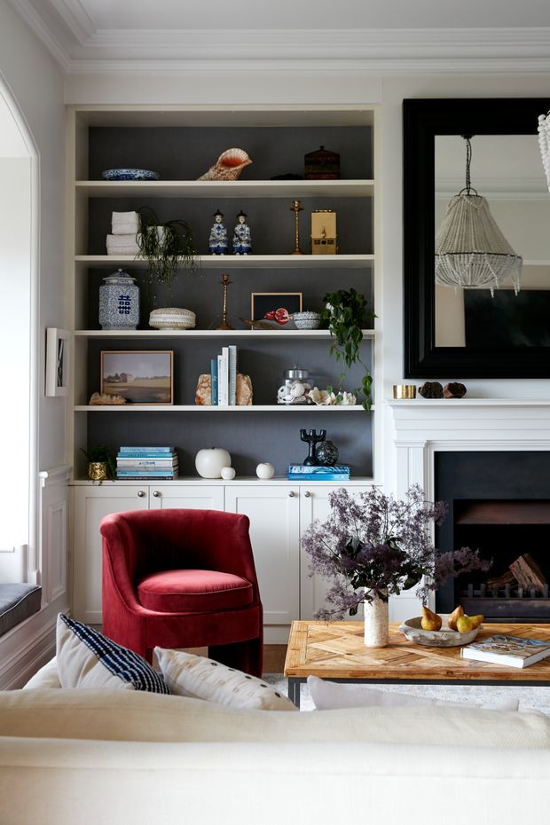 A refined living room with a built-in white display bookcase featuring dark charcoal backing, decorative objects, books and ceramics. The room includes a red upholstered armchair, a light neutral sofa, a timber coffee table, a classic white mantelpiece and black fireplace surround, framed artwork, and a sculptural pendant light. Detailed mouldings and cabinetry create a layered, elegant interior.
