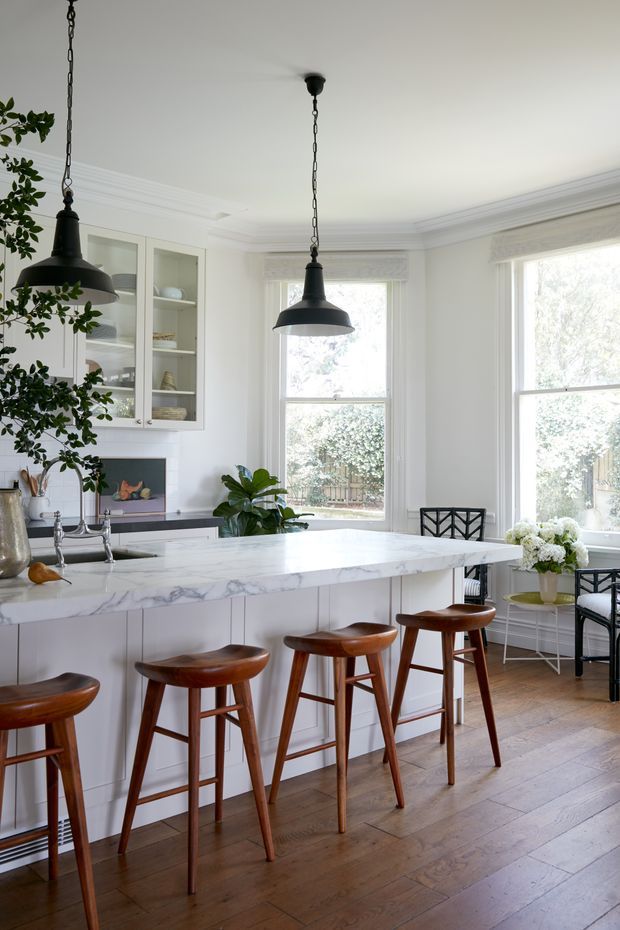 Bright, refined kitchen with a large white marble island and seating for four on warm wooden stools. White built-in glass-front cabinetry, timber flooring, bay windows with Roman shades, black pendant lights, and leafy plants create a classic yet contemporary interior.