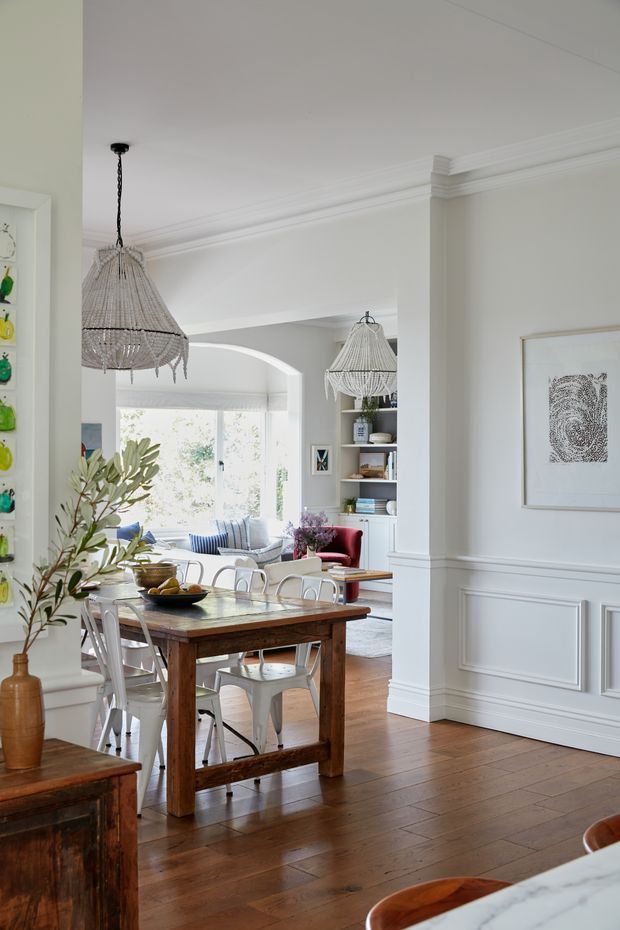 Bright, refined dining area in a period-style home, featuring a substantial timber dining table with metal chairs, white paneled walls with decorative mouldings, hardwood flooring, woven pendant lights, and an adjoining living area visible through a wide arched opening. Built-in shelving, artwork, and indoor greenery add warmth and character.