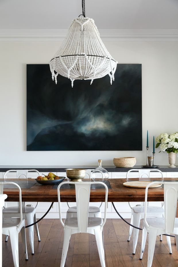 A refined dining room with a long rustic timber dining table surrounded by white metal chairs. A large dark abstract artwork hangs on a pale wall above a sideboard, complemented by a beaded chandelier, decorative vessels, candlesticks, and a floral arrangement. Timber flooring and detailed ceiling cornices add architectural character.