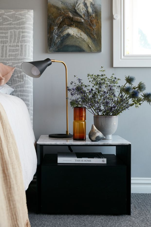 Close-up interior photograph of a styled bedside corner in a bedroom. A black bedside table with a pale stone-like top holds a modern black-and-brass lamp, amber glass vessels, a grey planter with leafy stems, and a small sculptural object. The scene includes a partially visible bed with white bedding and a textured grey upholstered headboard, a muted blue-grey wall with framed artwork, white window trim, and grey carpet.