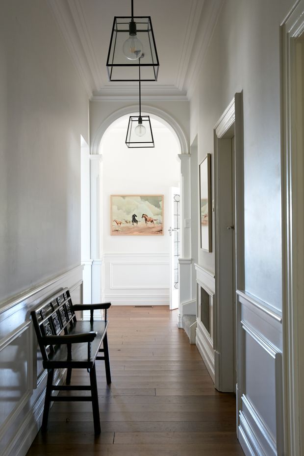 A bright, narrow residential hallway with pale walls, white decorative wainscoting and moulded architraves, a coffered ceiling, and a large arched opening at the far end. Two black geometric pendant lights hang along the corridor, while a dark timber bench sits against the left wall. Timber floorboards, framed artwork, and multiple doorways complete the refined traditional interior.