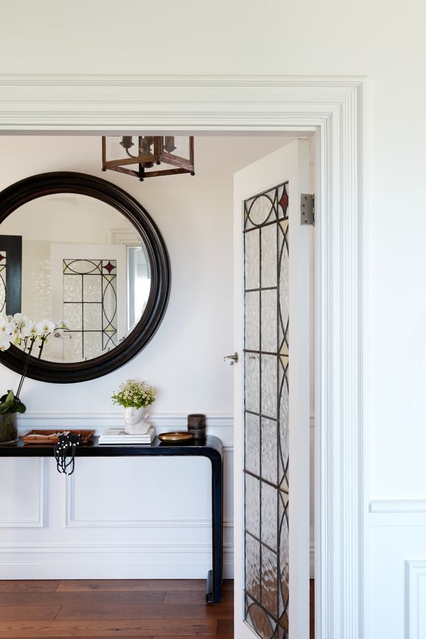 A refined traditional entryway with white painted wall panelling and elaborate layered trim around a doorway. A black console table sits beneath a large round dark wood-framed mirror, with floral arrangements and decorative objects. The open door features a distinctive leaded and stained-glass panel with geometric detailing, while warm timber flooring and a vintage pendant light add character.