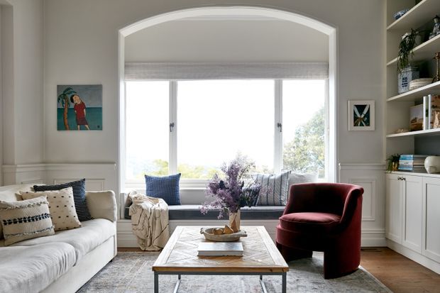 Bright, refined living room with a large arched bay window and built-in window seat, white paneled walls, open display shelving, a pale sofa layered with patterned cushions, a sculptural burgundy armchair, and a light timber coffee table over a muted area rug. The room has abundant natural light and an elegant, comfortable feel.