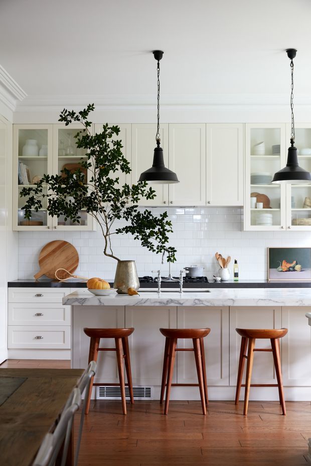 A bright, refined kitchen with white shaker-style cabinetry, glass-front display cupboards, a glossy white subway-tile splashback, dark benchtops, and a veined marble island. Three timber bar stools line the island, while black pendant lights hang from a detailed white ceiling. Warm timber flooring, open shelving, ceramic dishes, and a large indoor tree add character and natural warmth.