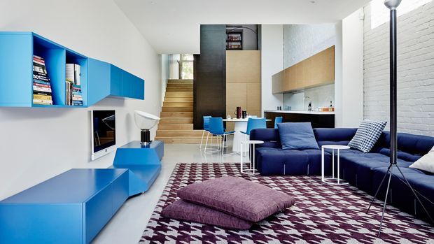 Bright open-plan contemporary living space with a vivid blue built-in media and shelving wall, blue sectional seating, patterned area rug, low floor seating, compact dining area, and a compact kitchen beneath a raised mezzanine or stair volume. White-painted brick and smooth white walls contrast with timber stair treads and warm wood cabinetry.