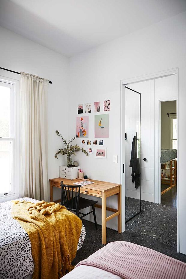 Bright, compact bedroom with white walls and ceiling, a timber writing desk and black chair, a mustard knitted throw over a patterned bed, and a second bed or upholstered bench in the foreground with a blush-pink cover. A full-height mirrored door reflects a bathroom or dressing area, while a small collage of artwork adds colour to the wall. The room has a simple, contemporary and playful feel.