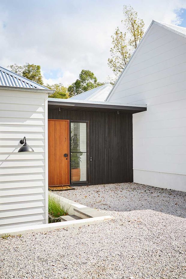 Exterior view of a contemporary residential entry courtyard. White horizontal weatherboard-clad buildings with steep gabled metal roofs frame a recessed entrance. The entry features a warm timber door, narrow glazed sidelight, dark vertically ribbed wall cladding, a black exterior wall light, and pale gravel landscaping with concrete edging.