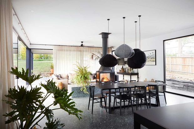 A bright, open-plan contemporary living and dining area with expansive glazing, sheer curtains, a dark dining table and chairs, and a suspended fireplace with a visible flame. Large leafy indoor plants, pendant lights, and views to a landscaped yard create a warm indoor-outdoor atmosphere. A kitchen island or benchtop is visible in the foreground.