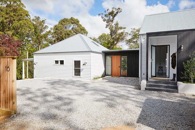 Exterior view of a contemporary residential property with a white weatherboard gabled structure connected to a darker charcoal-clad entry volume. The home has a corrugated metal roof, timber front door, gravel driveway, simple concrete steps, and mature trees surrounding the site.