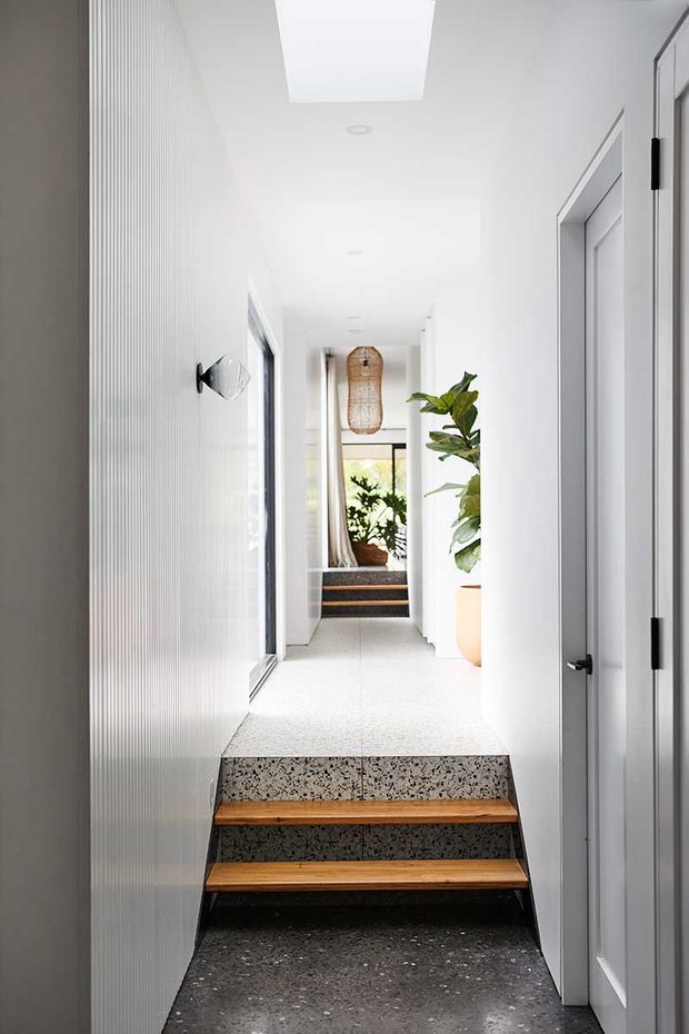 Bright contemporary residential hallway with a short flight of terrazzo and timber stairs, white vertically textured wall panelling, white doors and trim, skylight, recessed lighting, and indoor plants. The corridor continues to a glazed, plant-filled area at the far end.