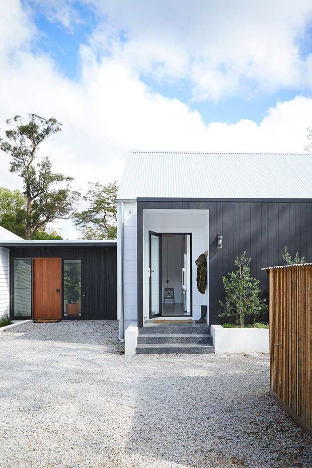 Exterior view of a contemporary single-storey home with a steep corrugated metal gable roof, dark vertical cladding, white-painted sections, and a glazed entry door reached by concrete steps. A detached or projecting dark-clad wing with a timber entry door sits to the left. The front yard is surfaced in light gravel and partly screened by a vertical timber fence, with mature trees around the property.