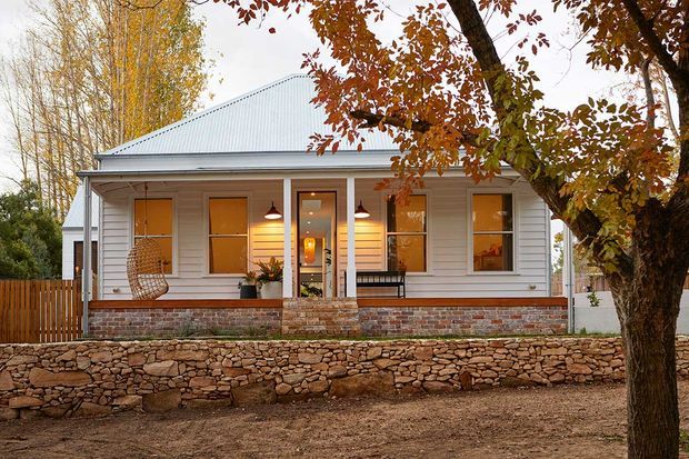 Front exterior view of a single-storey cottage-style home with pale horizontal weatherboard cladding, a simple pitched metal roof, warm timber-framed windows and doors, a covered entry veranda, and brick and rough-stone retaining walls. Mature deciduous trees frame the property.