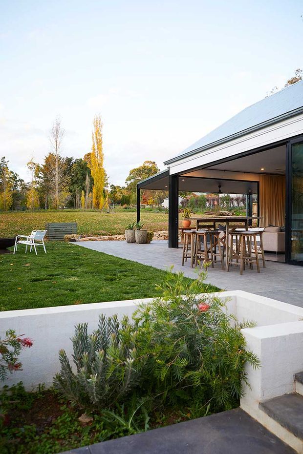 Contemporary single-storey home opening onto a landscaped garden, with a covered outdoor entertaining area featuring timber-clad walls, a dining table and stools, and large glazed openings. The low-pitched metal roof, dark slender framing, pale concrete terrace, raised white garden bed, and established lawn create a clean indoor-outdoor setting.