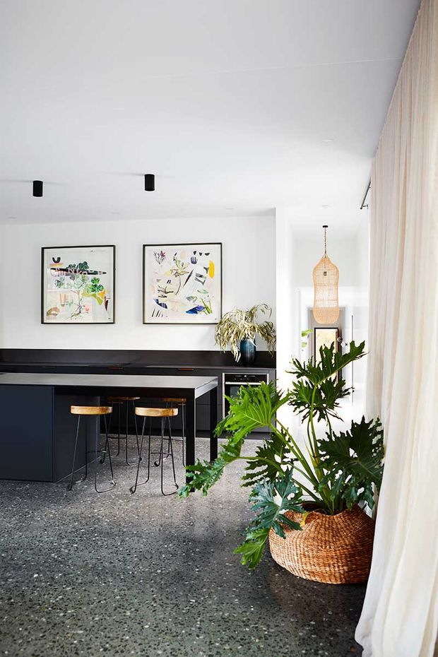 Bright contemporary kitchen and dining space with a dark island and cabinetry, terrazzo flooring, white walls and ceiling, framed botanical artwork, timber bar stools, woven pendant lighting, and large potted tropical plants beside sheer curtains.