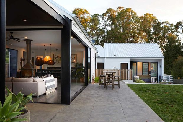 Contemporary single-storey home with a dark-framed glazed living area opening onto a broad paved terrace and lawn. The house has a crisp white exterior, corrugated metal pitched roof, and a connected rear wing. Through the glazing, an open-plan living and dining area is visible with a sofa, pendant lights, exposed structural elements, and indoor planting. Mature trees form the background.