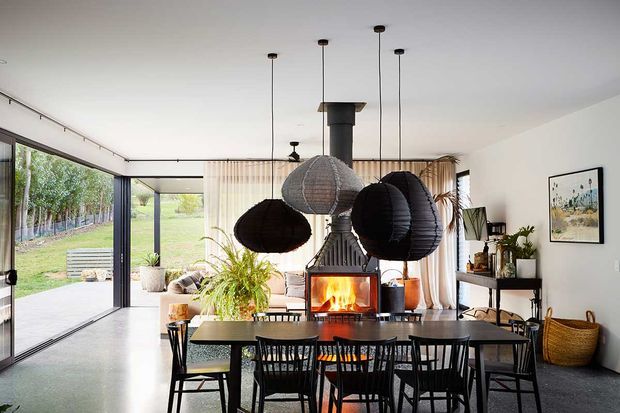 Bright open-plan dining and living space with a large black dining table and spindle-back chairs positioned in front of a central wood-burning fireplace. Oversized sculptural pendant lights hang from the ceiling, while full-height glazing and sliding doors open to a grassy outdoor setting. The room includes light curtains, indoor plants, a sideboard, and modern artwork.
