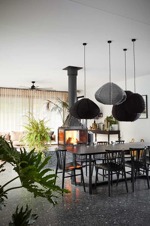 A stylish open-plan dining and living area centered around a contemporary suspended fireplace with a visible fire. A dark dining table and black spindle-back chairs sit beneath several oversized woven pendant lights. The room features polished dark terrazzo flooring, pale walls and curtains, abundant indoor greenery, a ceiling fan, and a low sideboard with decorative objects. A lounge area is visible behind the dining zone.