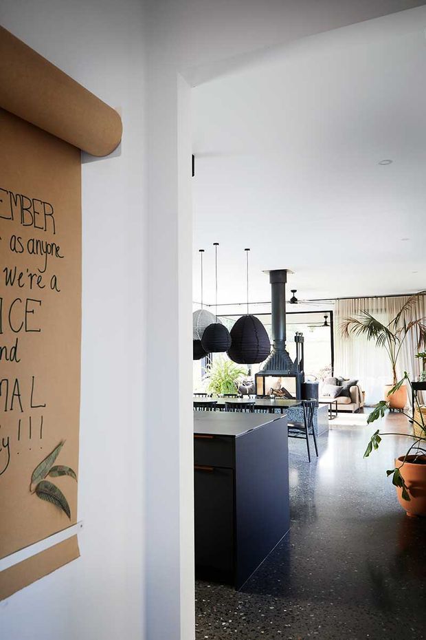 View through a white-framed opening into an open-plan contemporary living and kitchen area. The space features a dark polished terrazzo floor, black kitchen cabinetry, pendant lights, a freestanding wood-burning stove, exposed dark ceiling fans, large glazing with sheer curtains, and abundant indoor plants. A large illustrated wall panel is visible in the foreground.